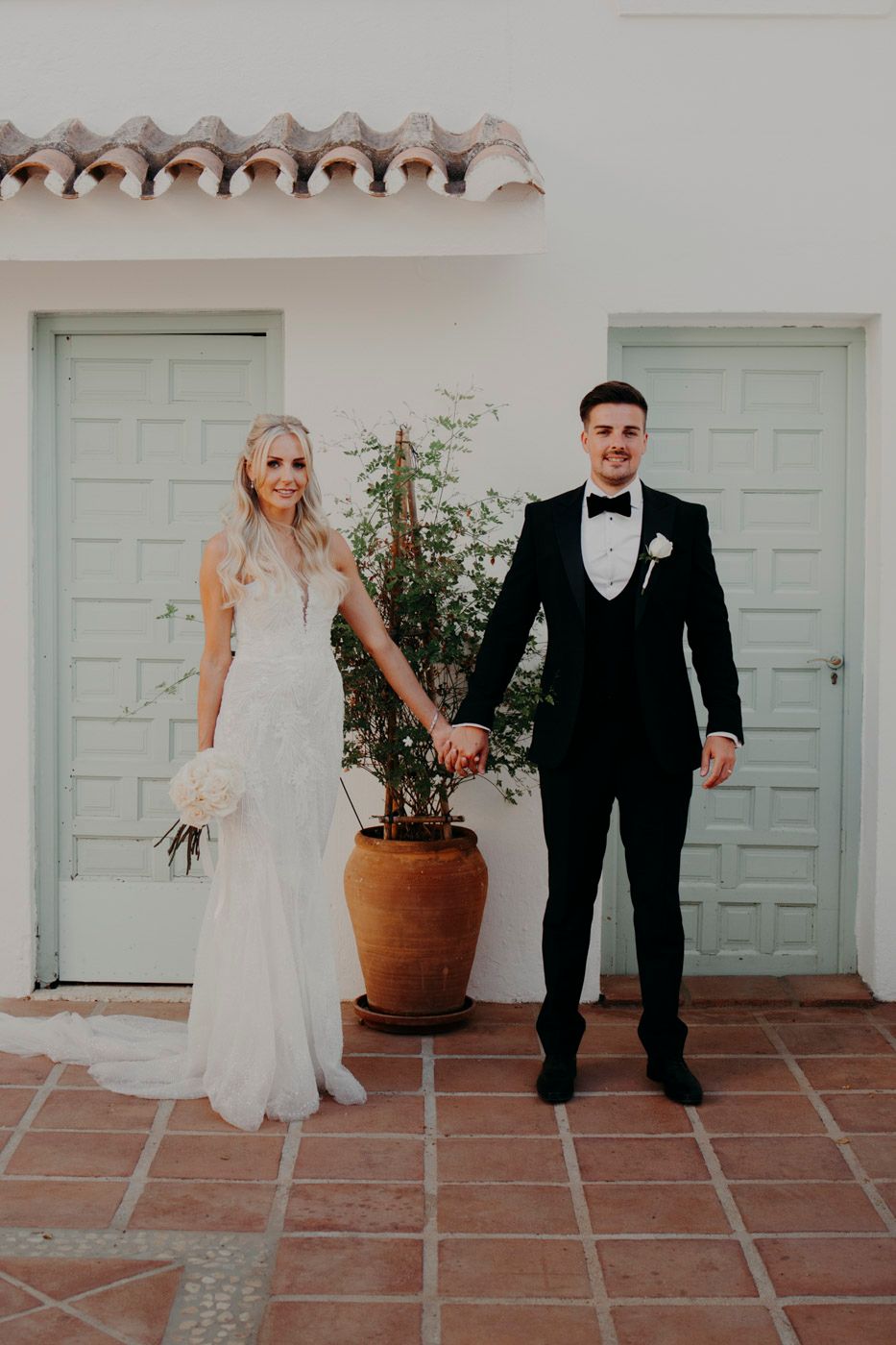 A wedding couple poses on terra cotta tiles in front of white Spanish-style doors.