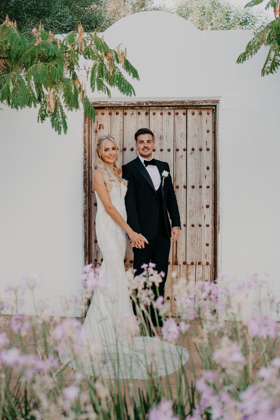 A romantic wedding couple pose in front of a rustic wooden door surrounded by delicate purple flowers in bloom.