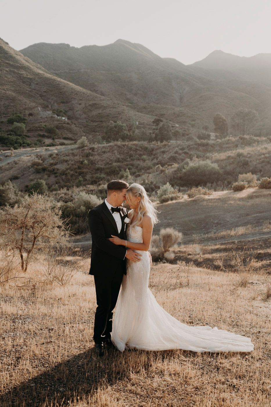 A couple shares an intimate moment in their wedding attire against a backdrop of golden mountains at sunset.
