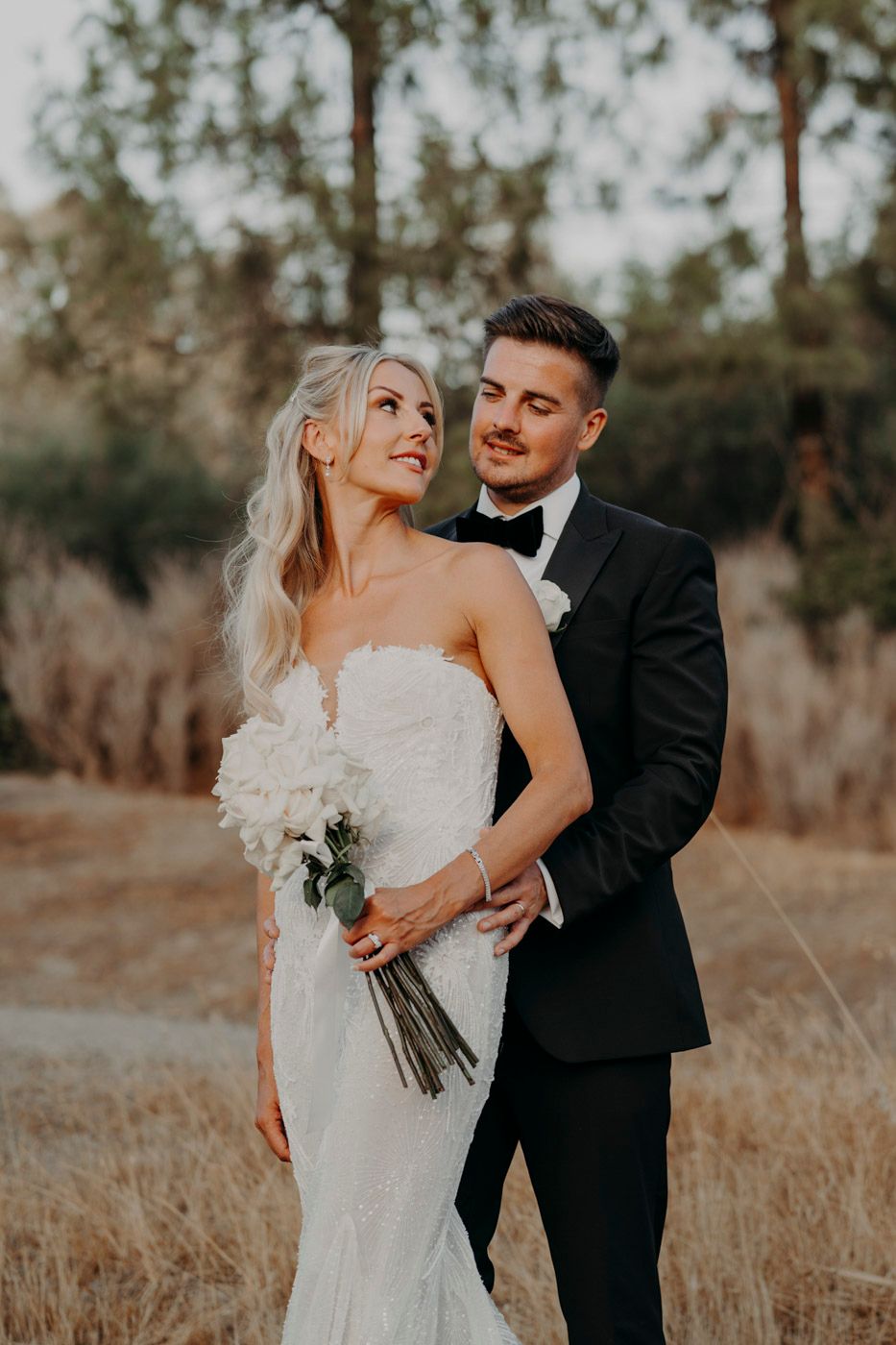 A romantic outdoor wedding portrait of a couple in formal attire embracing in a golden field at sunset.