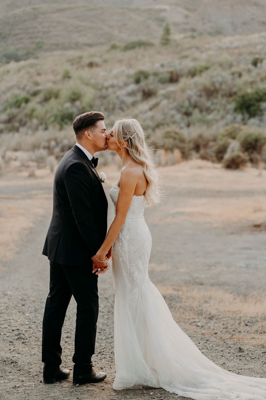 A romantic couple shares a kiss on a desert hillside path with the bride wearing a flowing white gown.