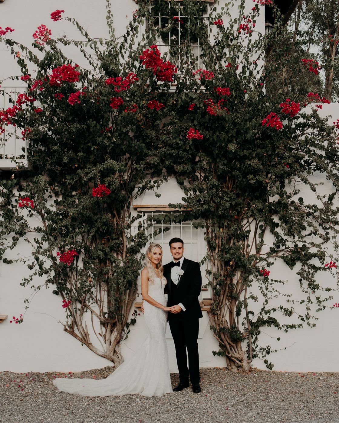 A couple stands beneath a beautiful archway of red flowering vines against a white wall.