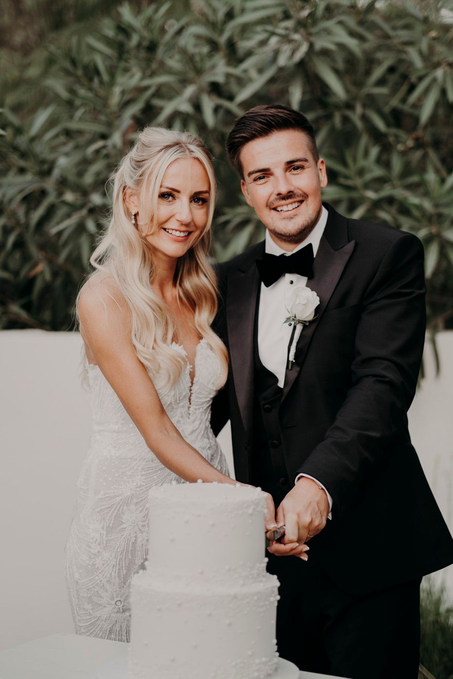 Newlywed couple smiling together while cutting their elegant white wedding cake in an outdoor garden setting.