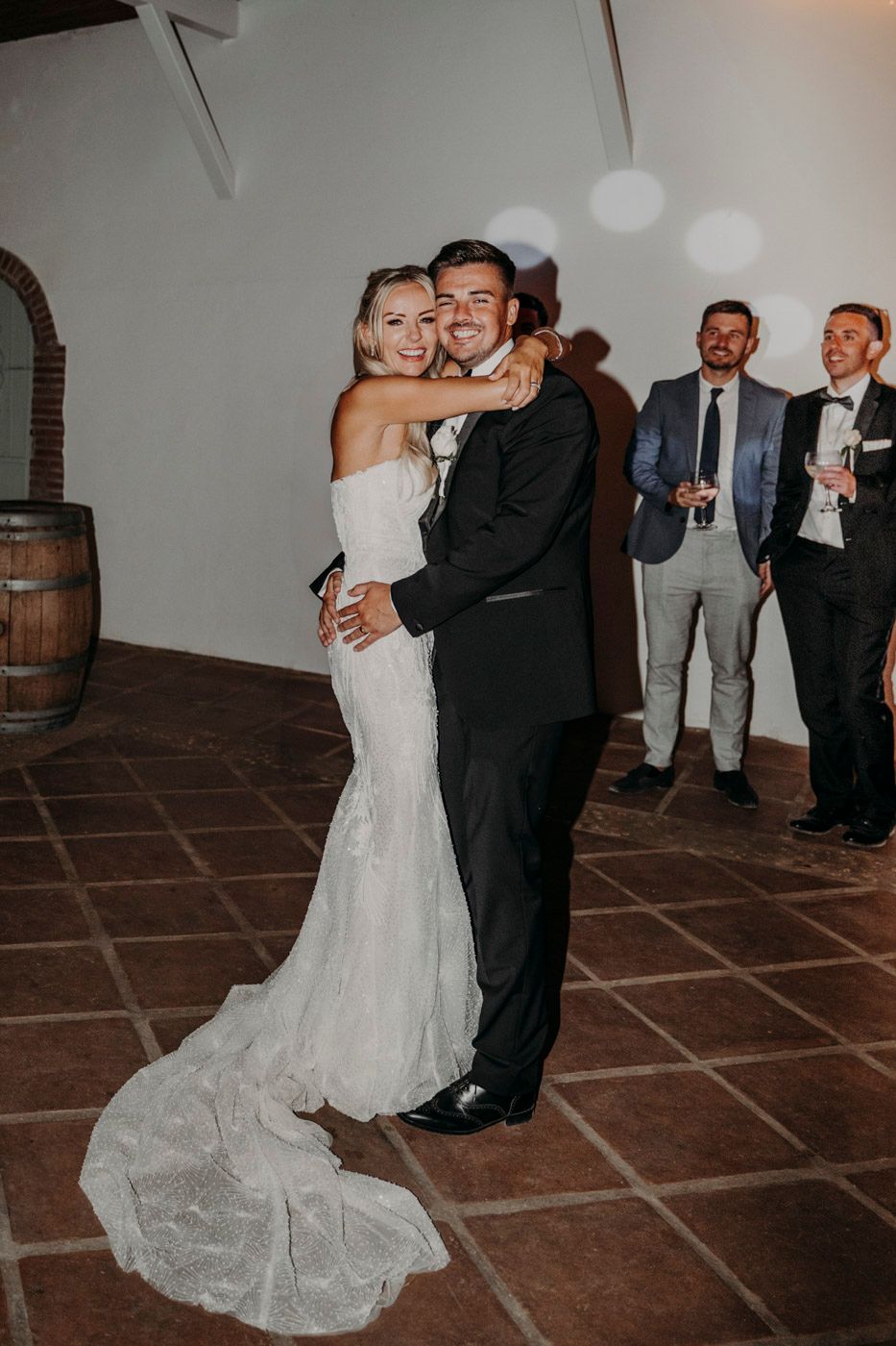Newlyweds share their first dance at rustic venue with wine barrel and guests watching in background.