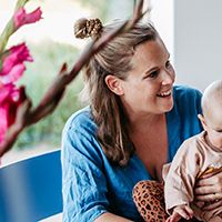 Frau in blauem Oberteil lächelt und hält ein Baby. Im Vordergrund sind unscharfe rosa Blumen zu sehen.