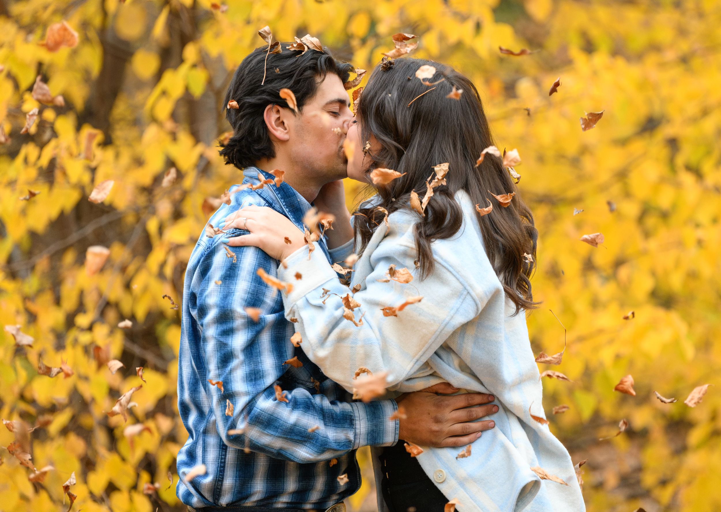 A couple shares a romantic kiss surrounded by golden autumn leaves in a park setting.