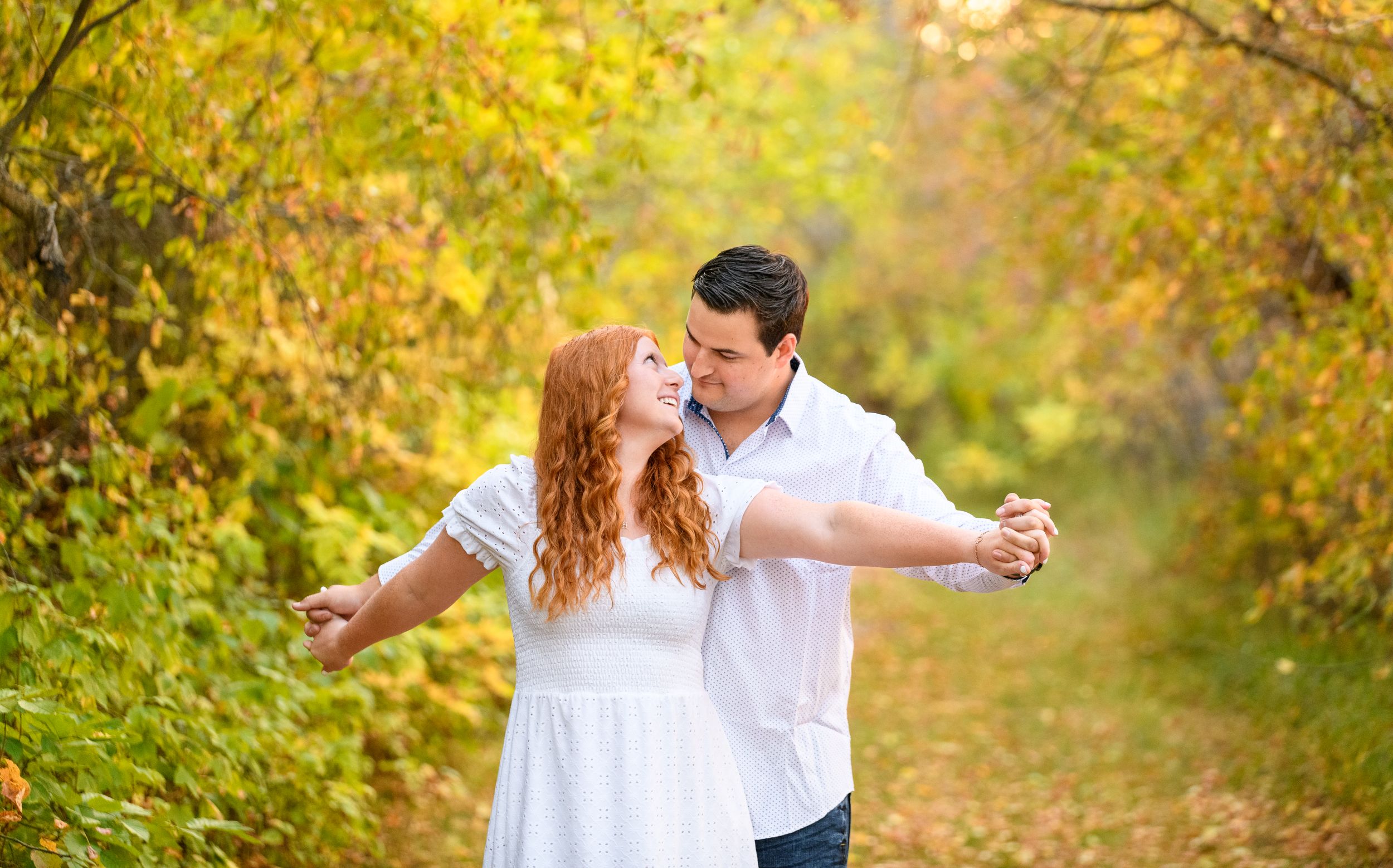 A couple in white outfits dances together on a wooded path during autumn with golden foliage in the background.