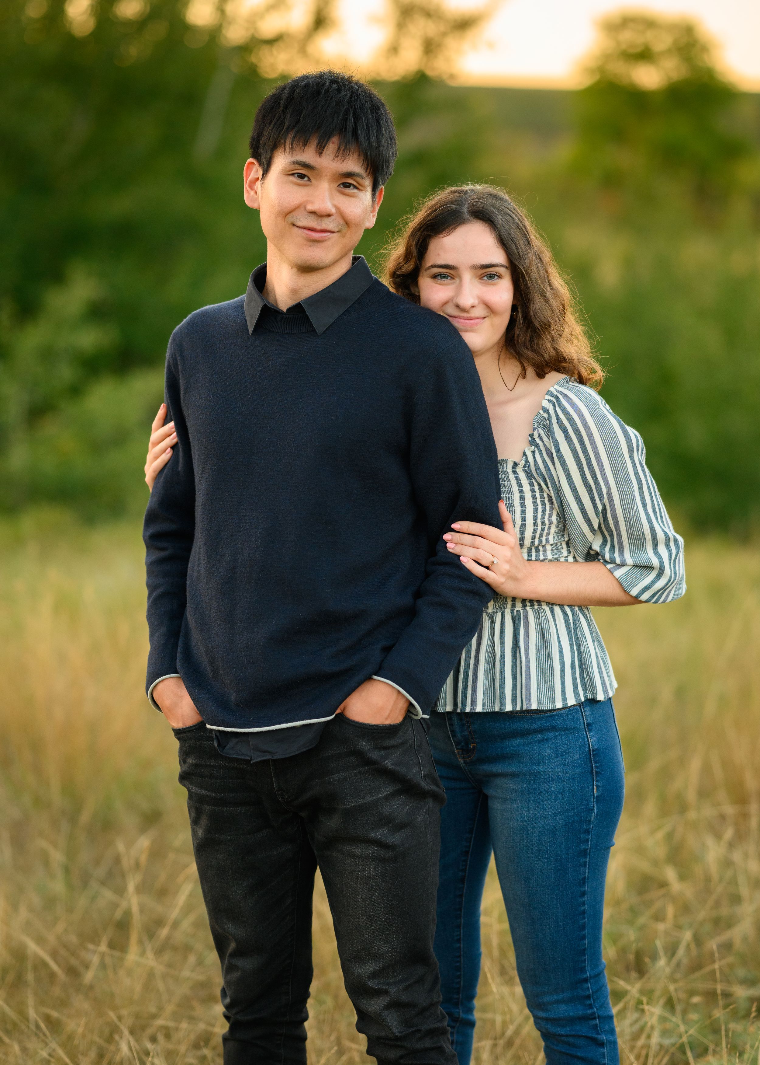 A couple smiles together in a sunny field during golden hour, with one wearing a black sweater and the other a striped top.