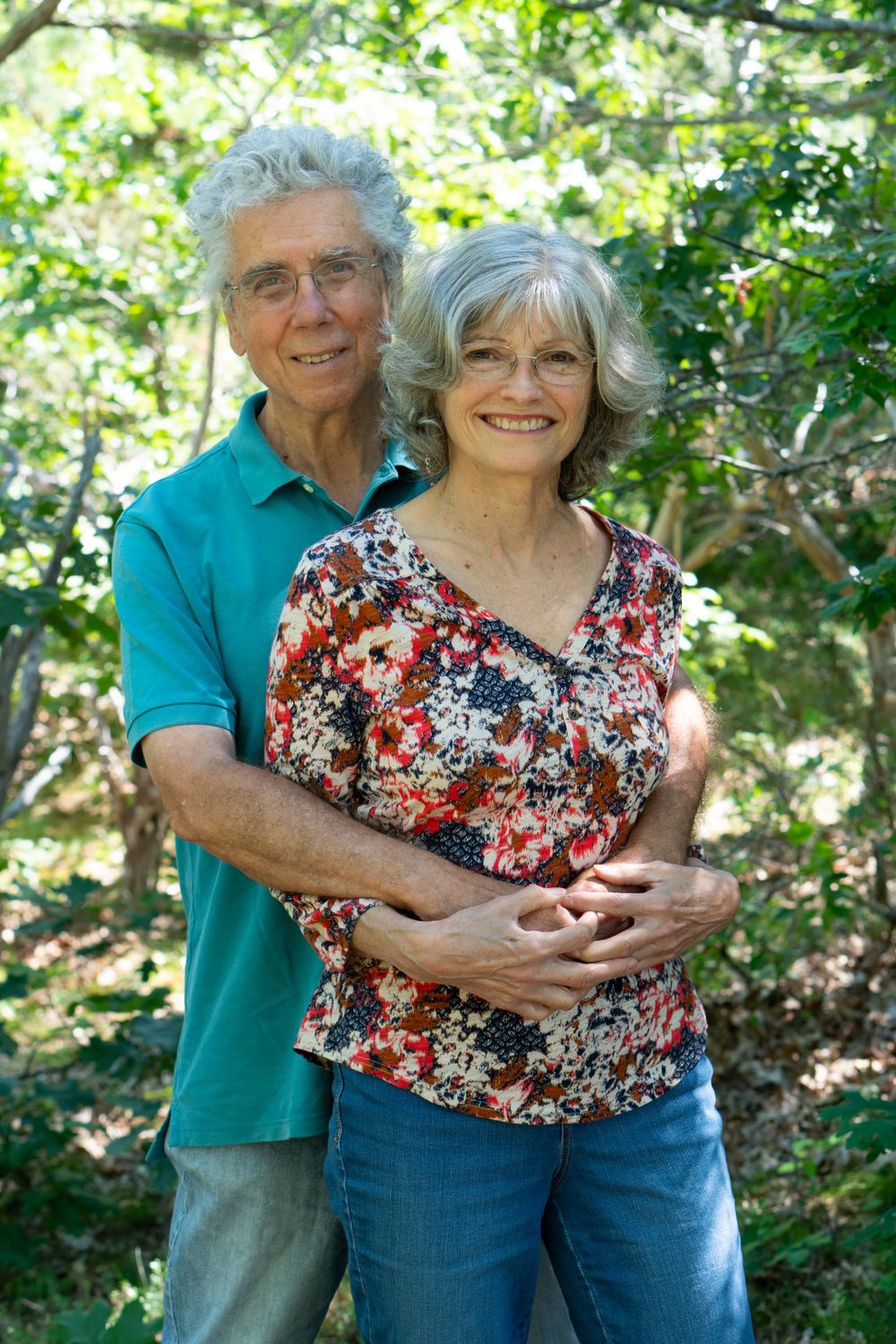 Senior couple embracing outdoors in casual clothing among green foliage.