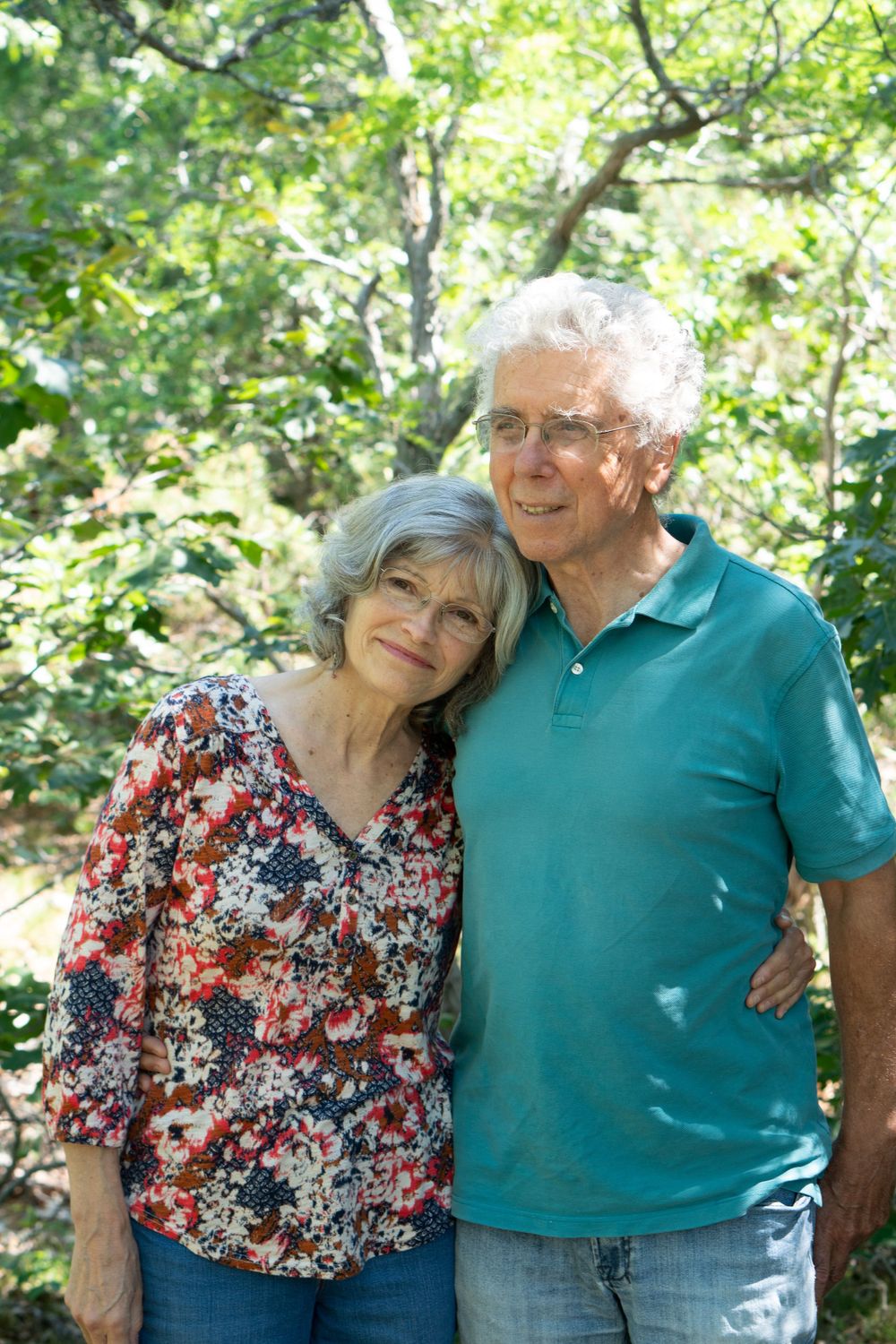 Mature couple in casual wear smiling together in sunny outdoor setting, with green trees in the background.