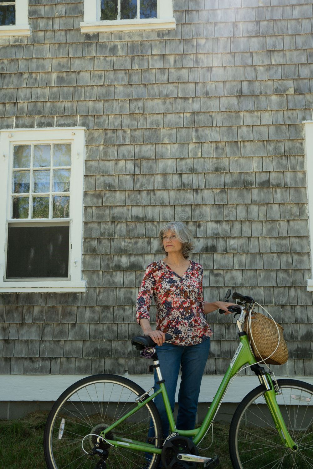 Woman with silver hair standing next to green bicycle against weathered wooden building.