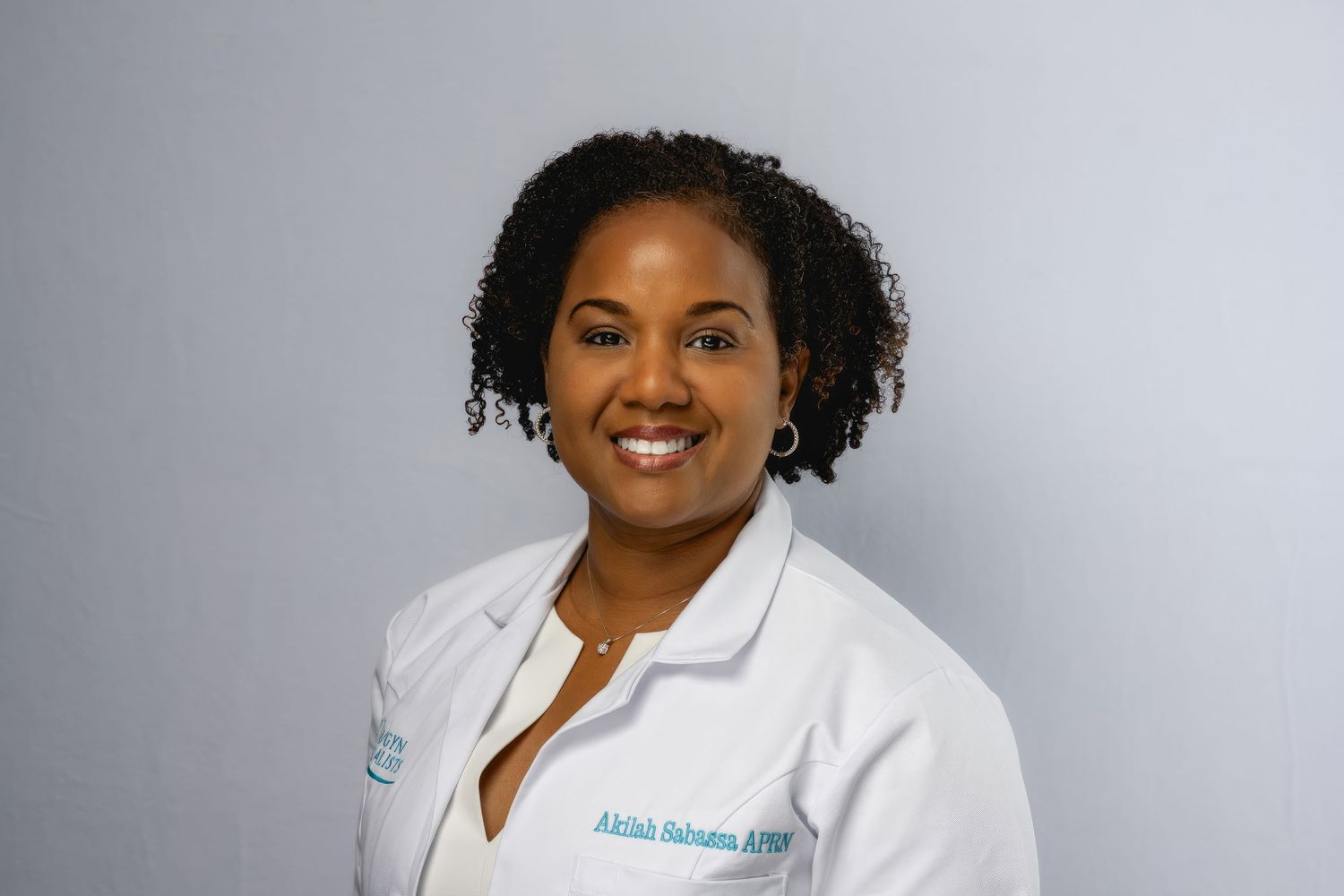 Medical professional in white coat smiles warmly against a gray studio background.