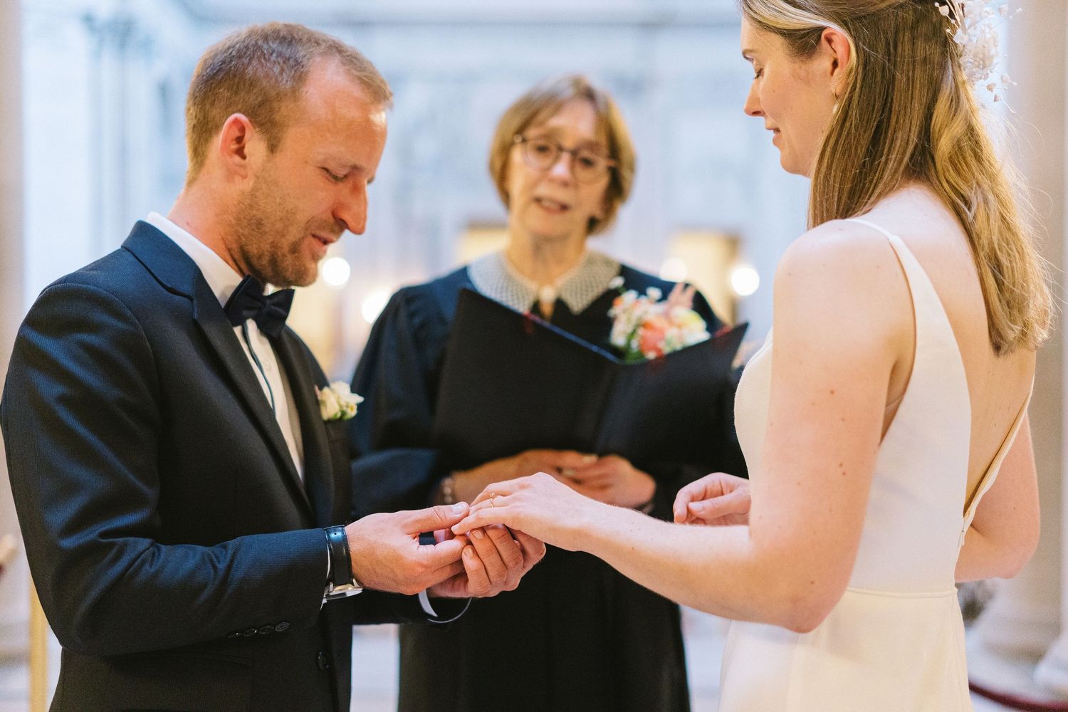 A couple exchanges rings during an indoor wedding ceremony with an officiant presiding.