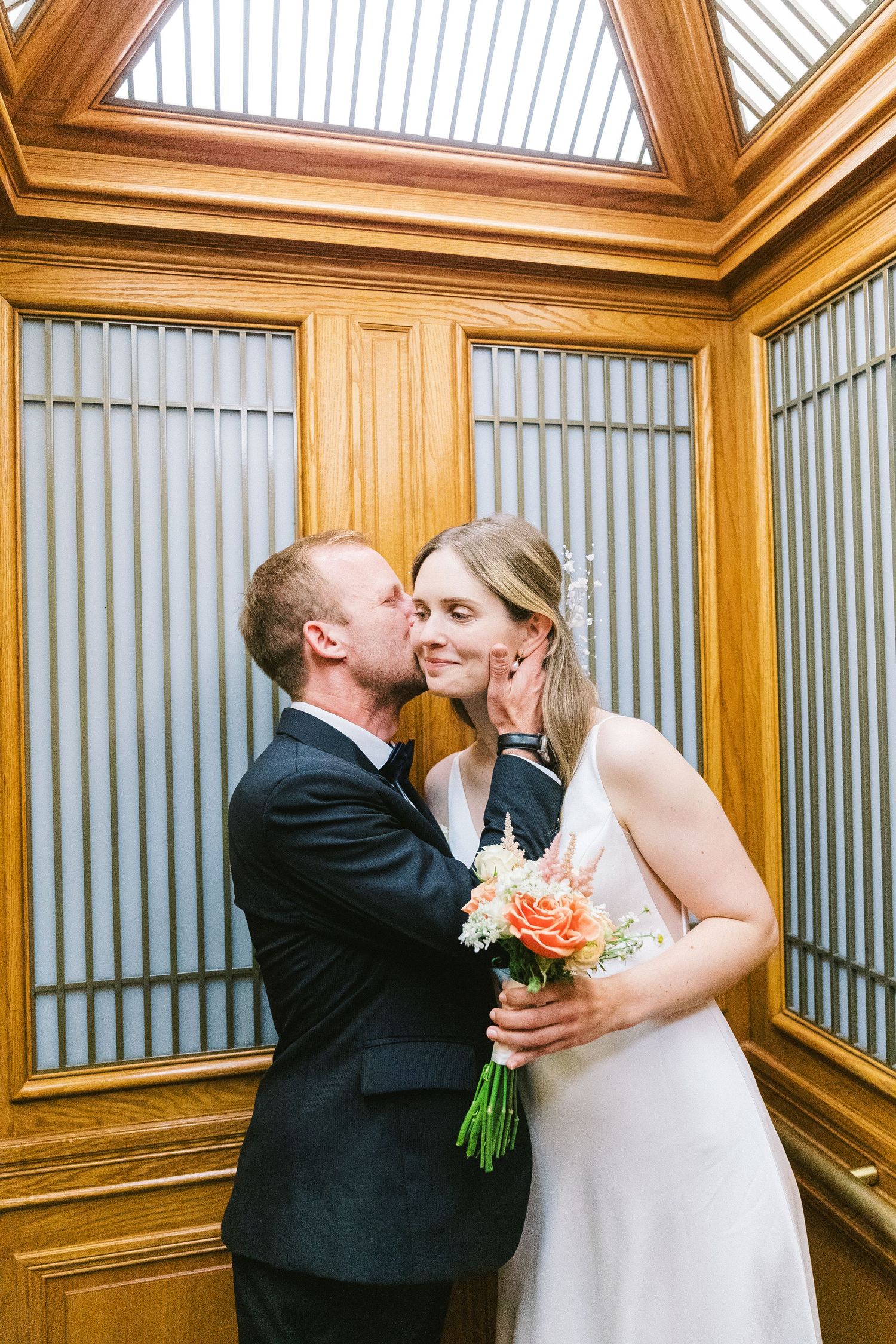 A romantic moment captured in an elegant wooden elevator with peach floral bouquet and formal wedding attire.