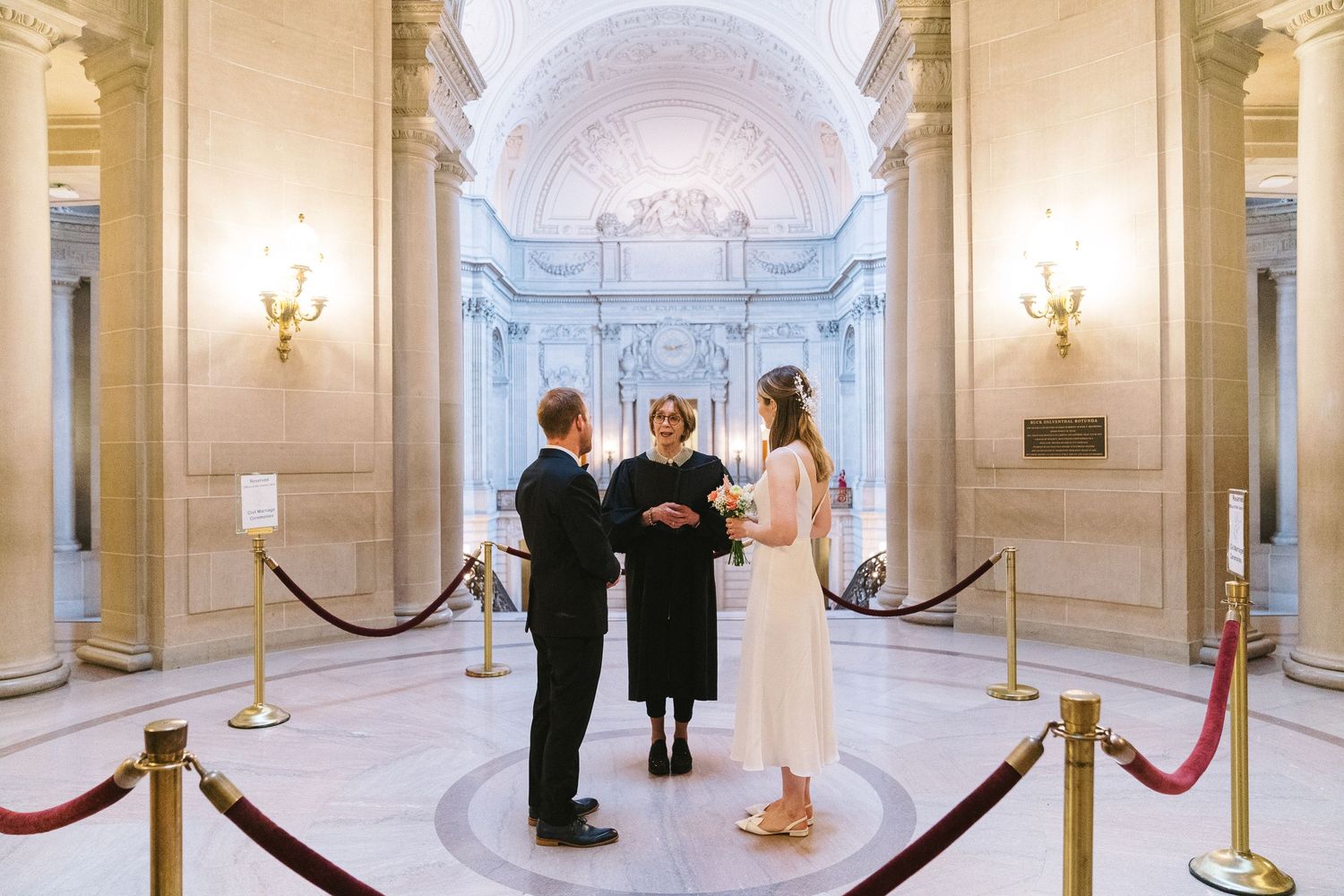 A wedding ceremony takes place inside a grand marble hallway with vaulted ceilings and elegant wall sconces.