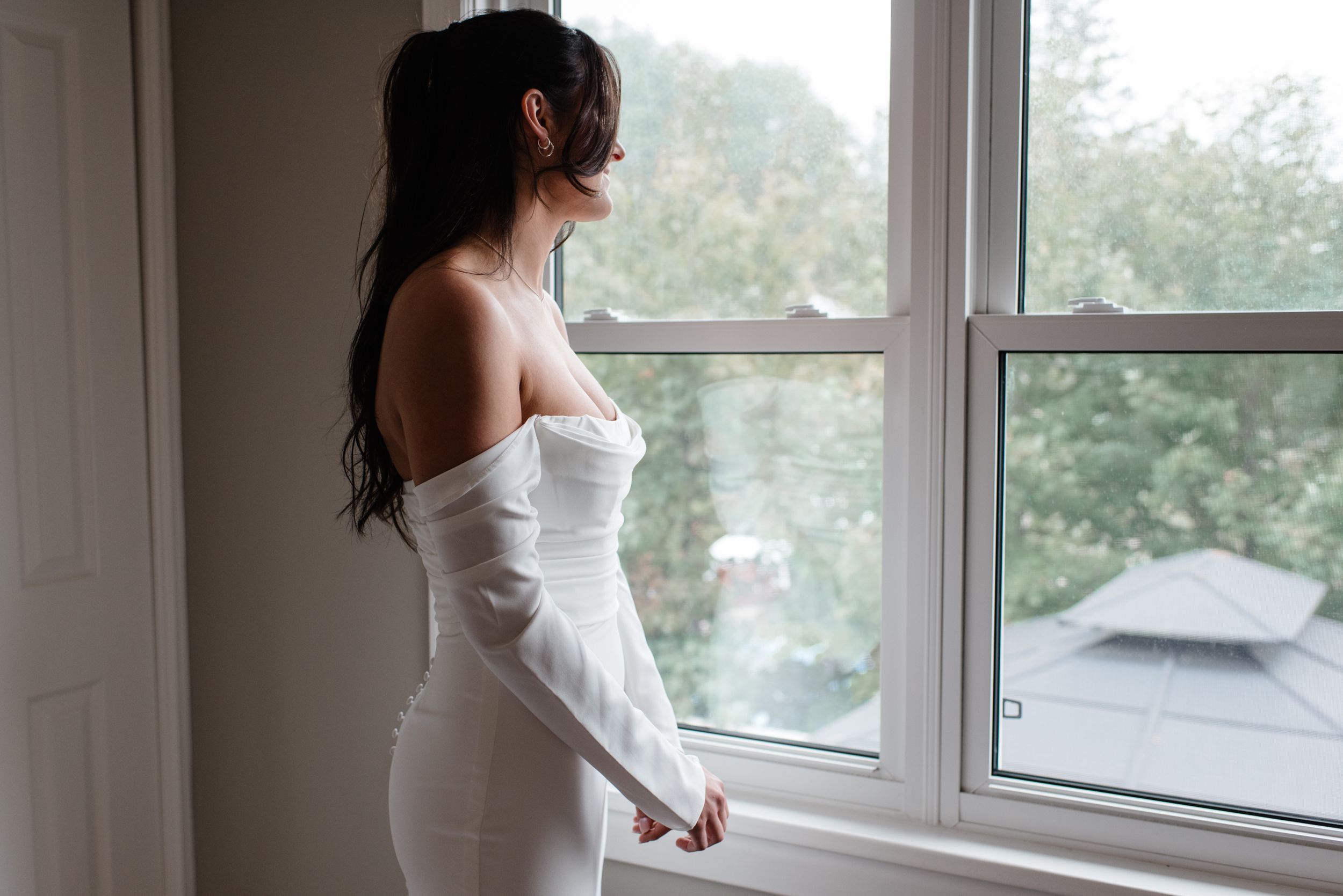 A person in an elegant white off-shoulder dress gazes out a window in soft natural lighting.