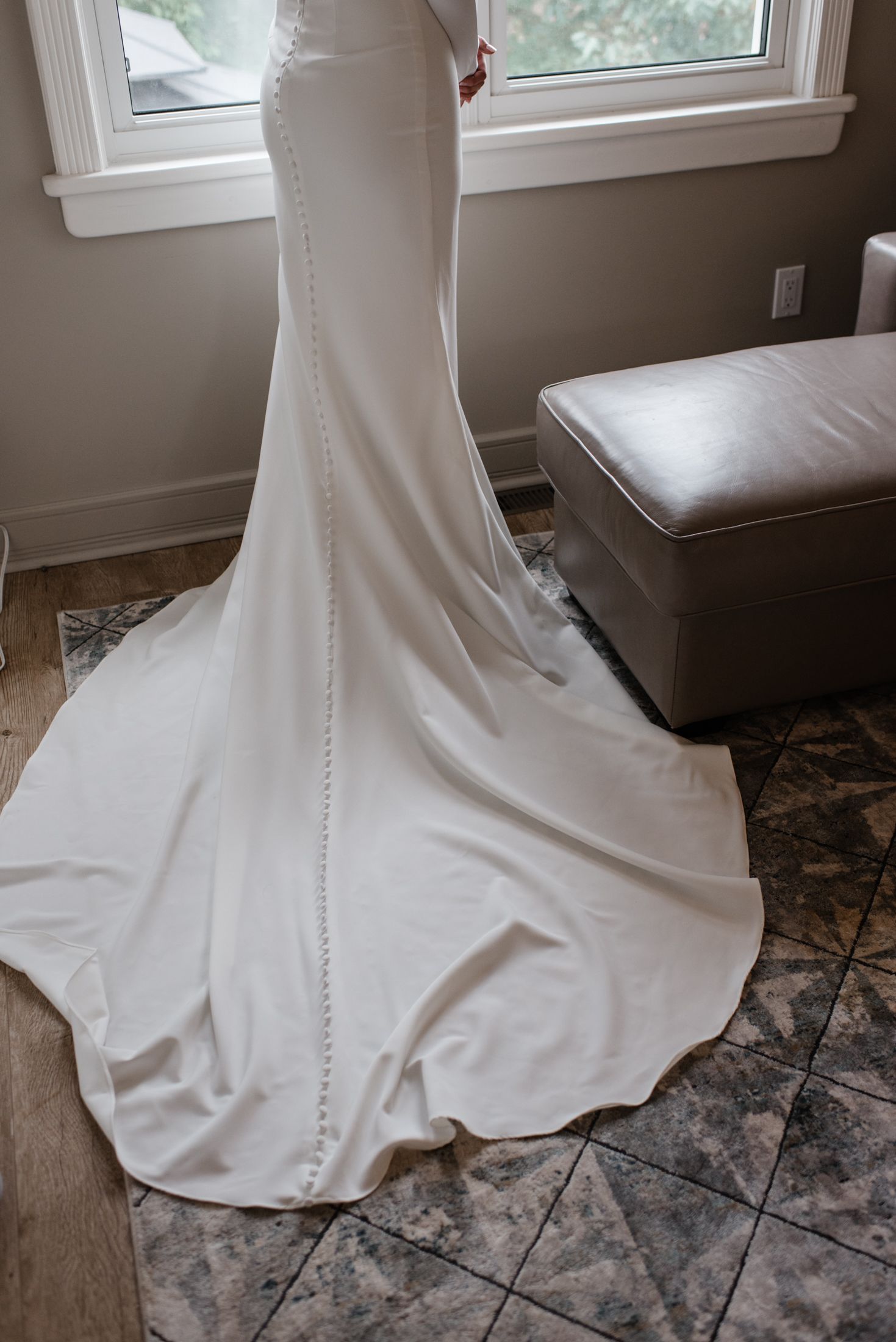 The train of an elegant white wedding dress with delicate button details flows across gray tile flooring.