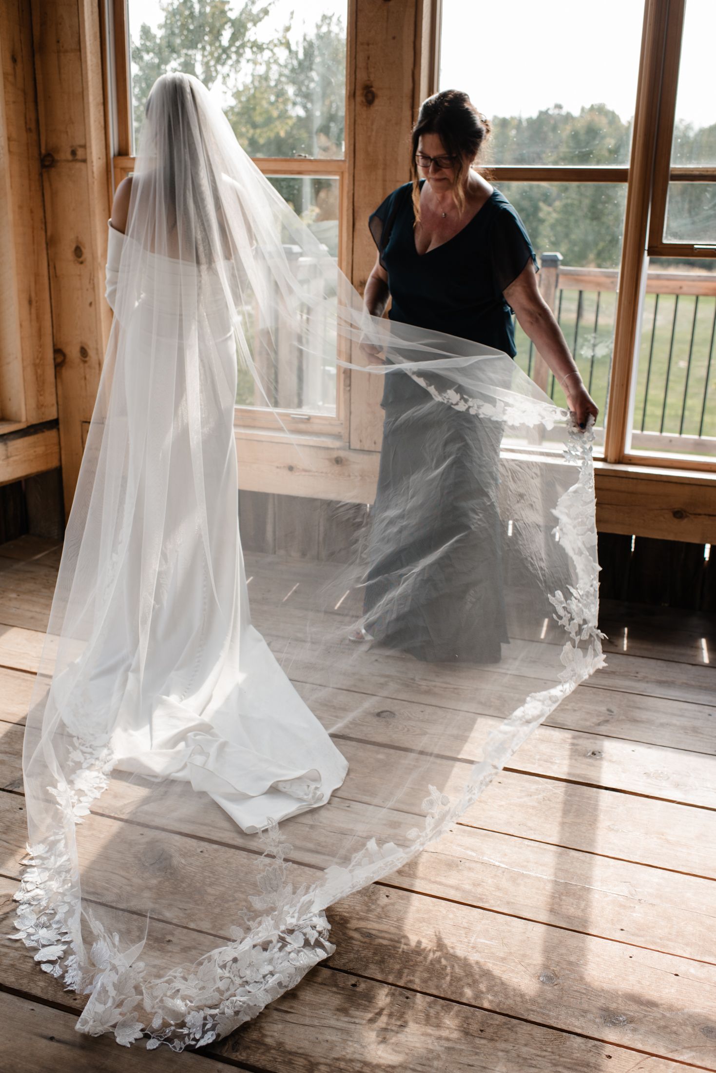 Two people adjust a flowing white lace wedding veil on a rustic wooden floor with large windows and natural lighting.