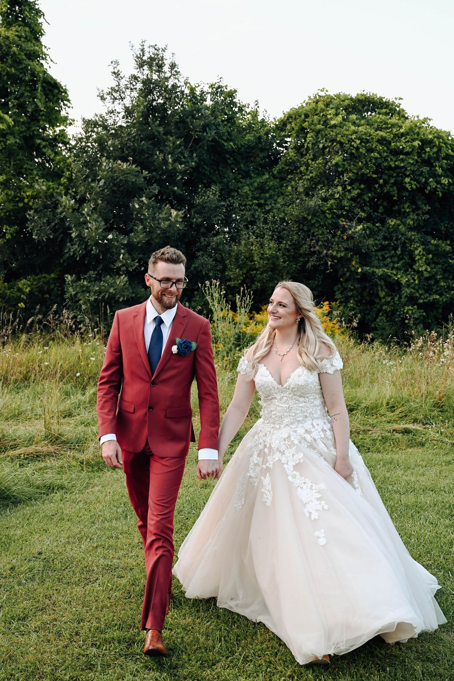 A couple walks on grass wearing formal wedding attire with a red suit and white lace gown against greenery.