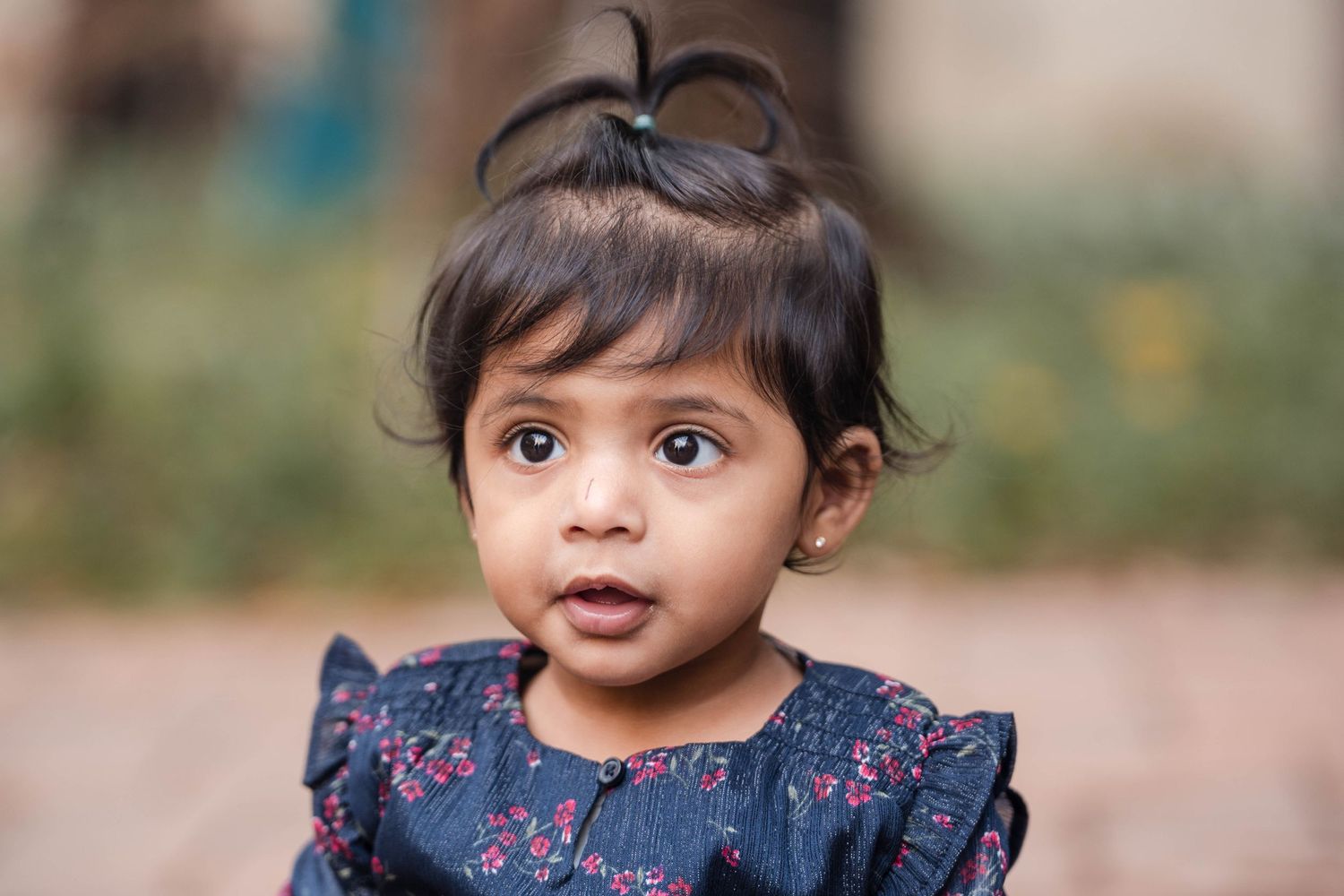 Adorable Indian toddler girl with dark eyes and hair in a ponytail wearing a floral dress outdoors.