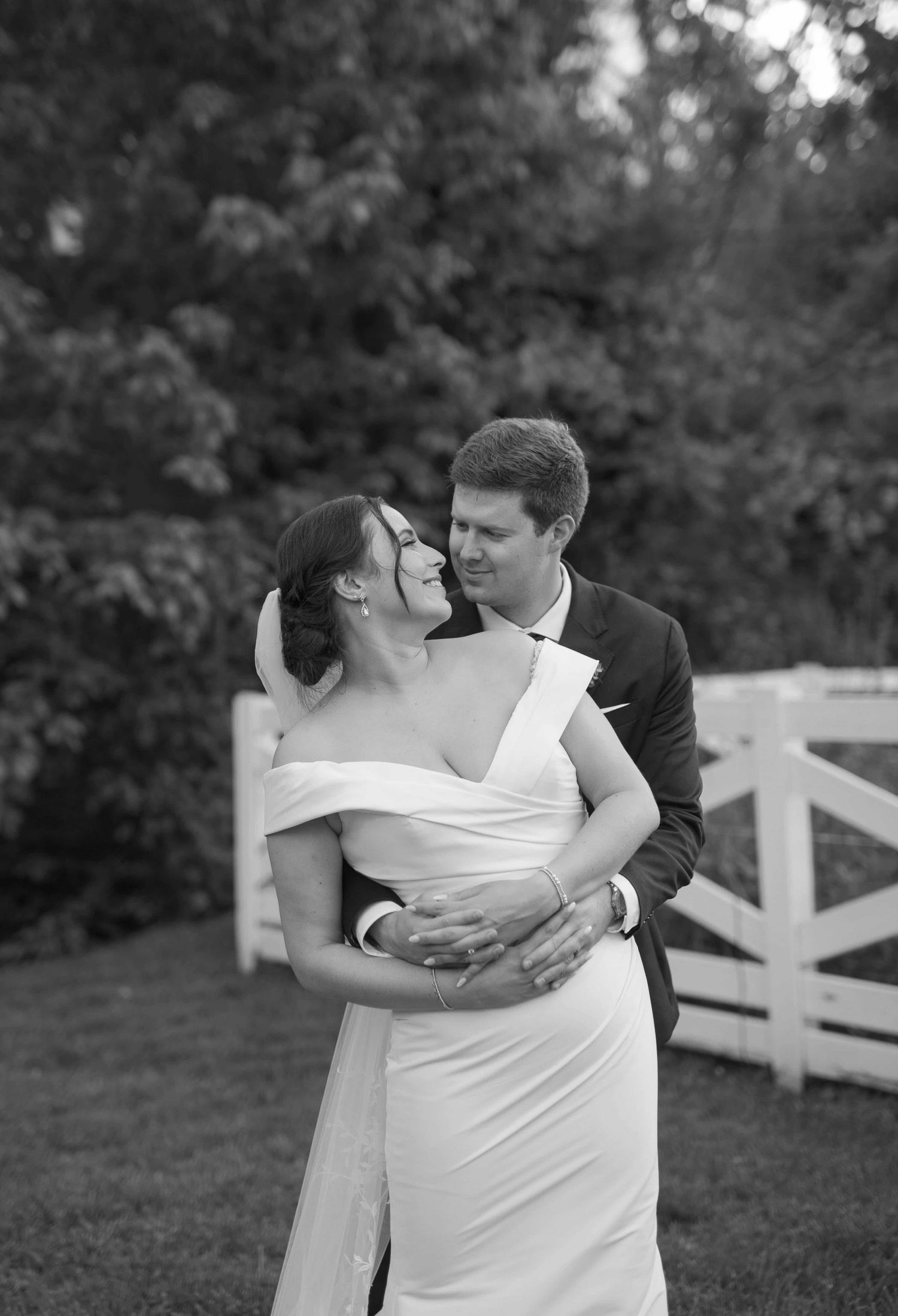 A romantic black and white wedding photo shows a couple embracing outdoors with a white fence in the background.