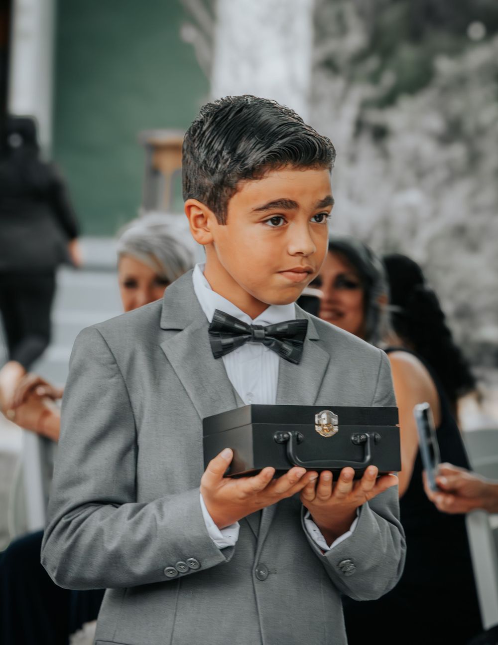 Young attendant in a gray suit and bowtie holds a decorative box during a formal event.
