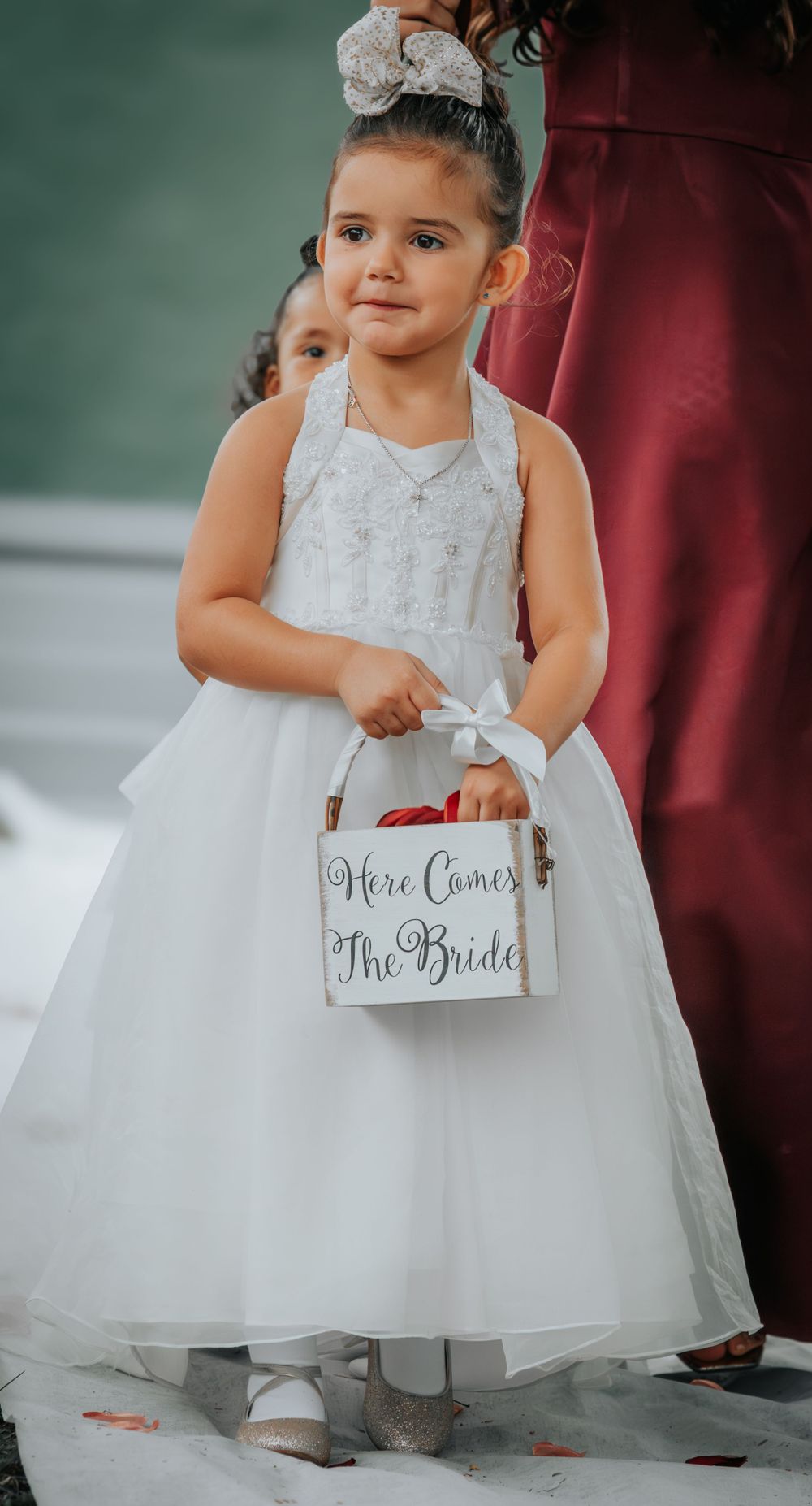 Flower girl in white tulle dress holds 'Here Comes The Bride' sign at wedding ceremony.