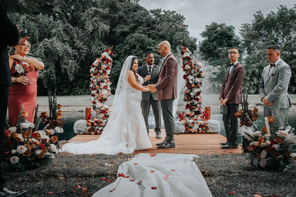 Outdoor wedding ceremony with floral arches and rose petals lining aisle as couple exchanges vows on wooden platform.