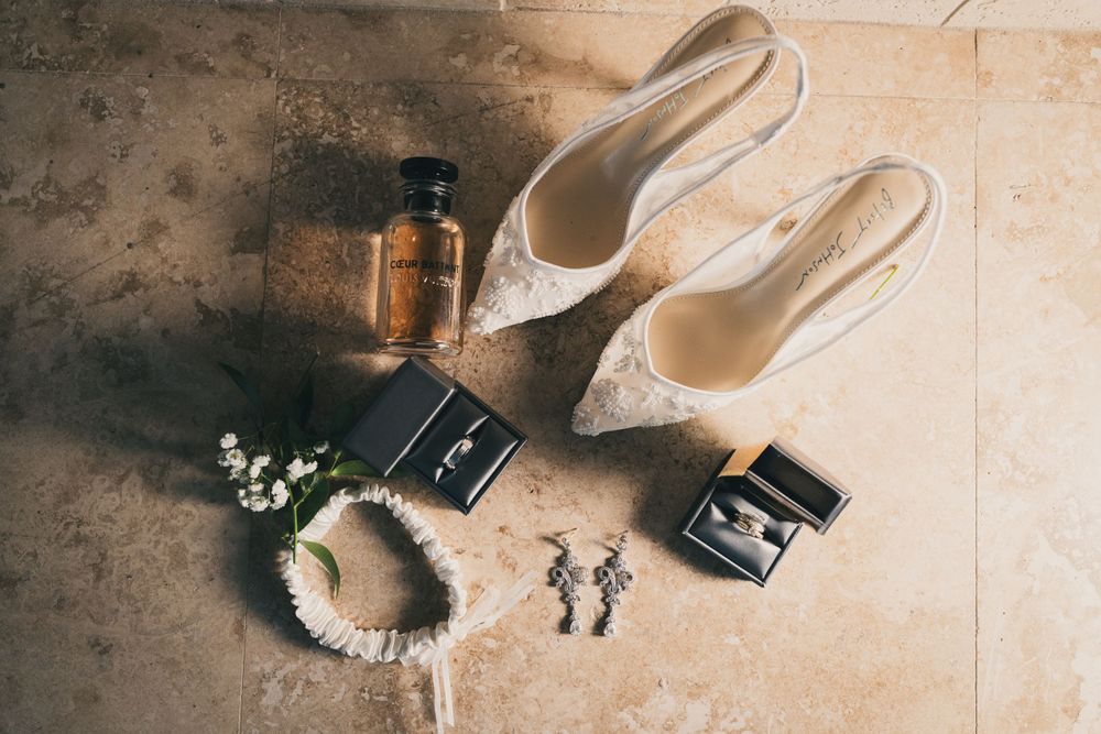 Wedding accessories including white lace heels, perfume bottles, earrings, and a white floral headpiece on a textured surface.