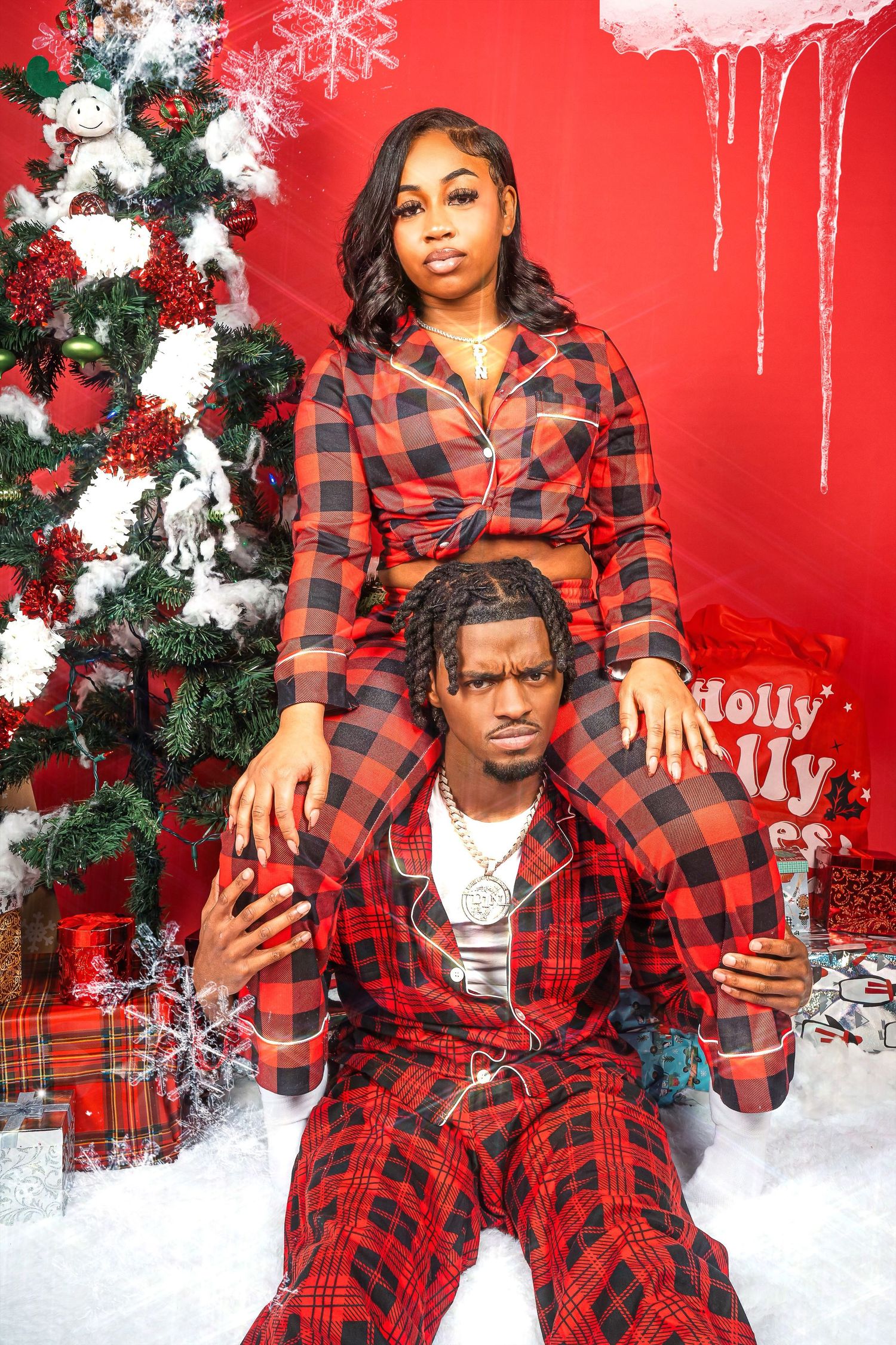 A festive holiday photo with two people in matching red plaid pajamas posing by a decorated Christmas tree against a red backdrop.