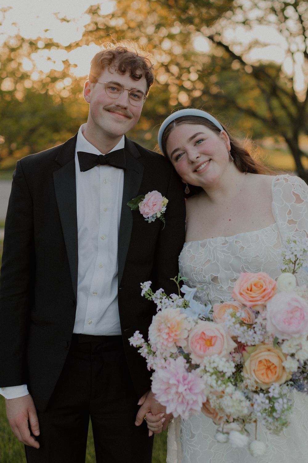 Romantic wedding couple poses with a beautiful bouquet of peach and pink flowers against an autumn backdrop.