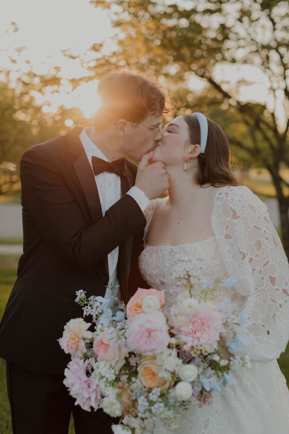 Newlyweds share a romantic kiss at sunset surrounded by pink and white flowers.