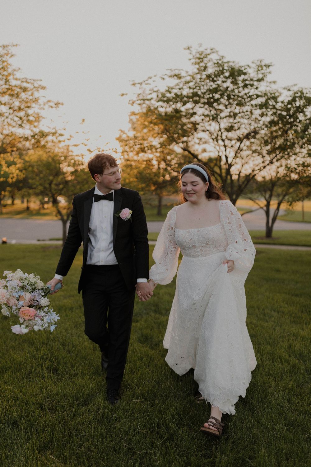 A couple walks hand in hand on grass during sunset, with the bride wearing a lace dress and groom in classic tuxedo.