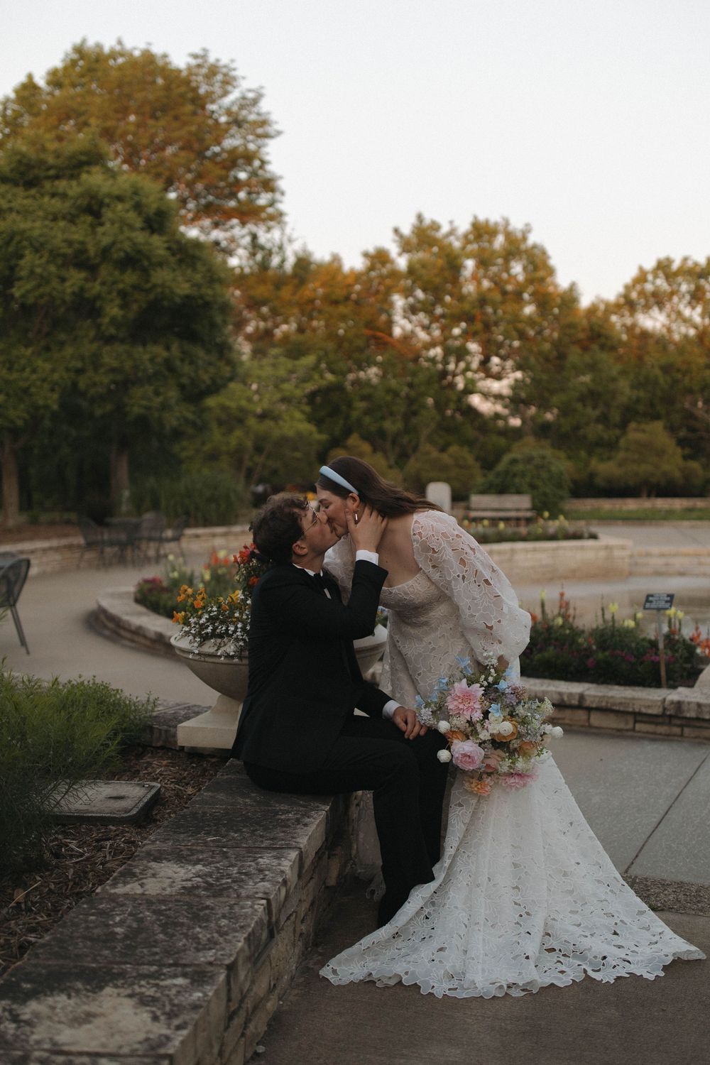 A romantic wedding moment captured in a garden setting with autumn trees in the background.