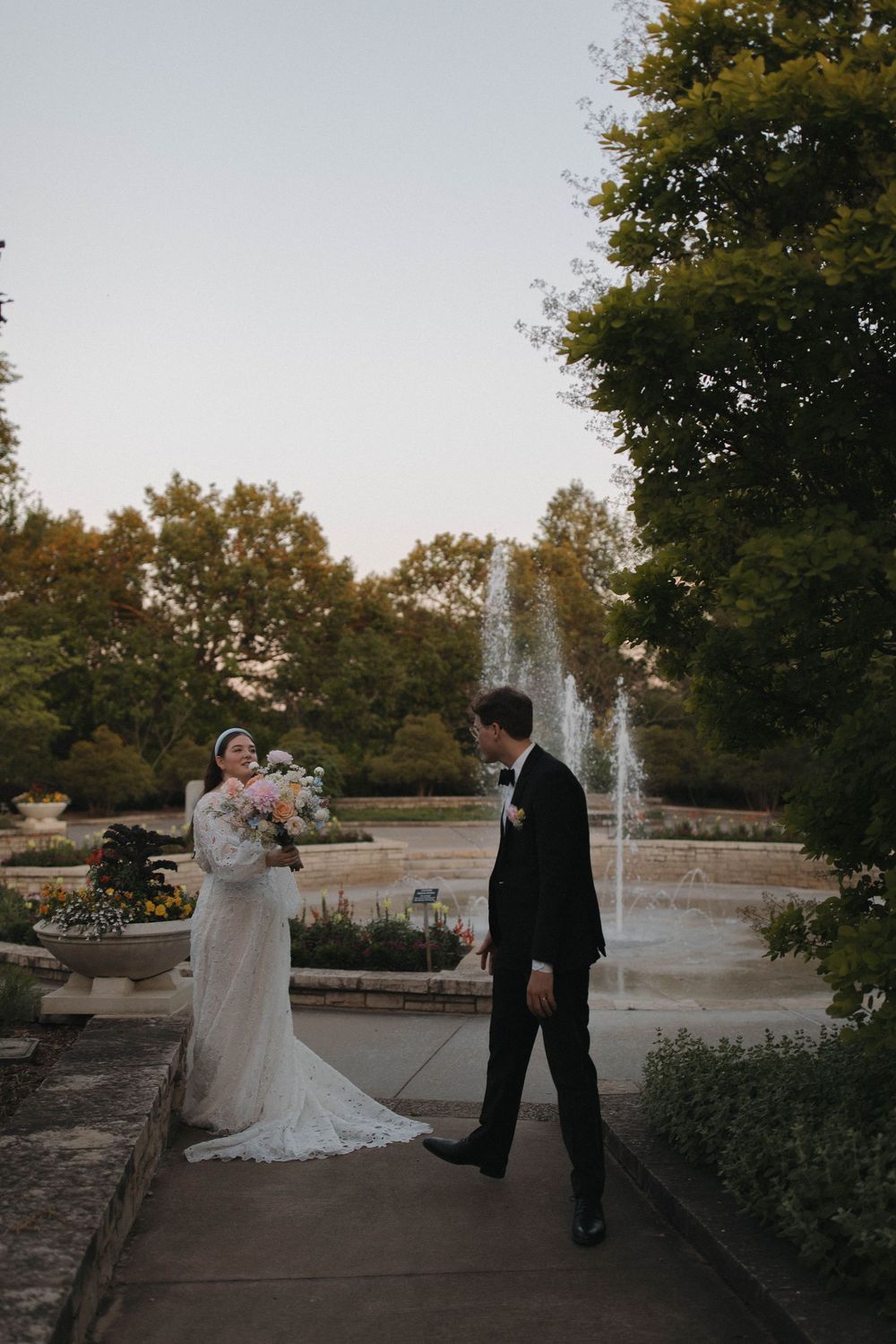 Wedding couple poses together near fountain and trees in formal garden setting at dusk.