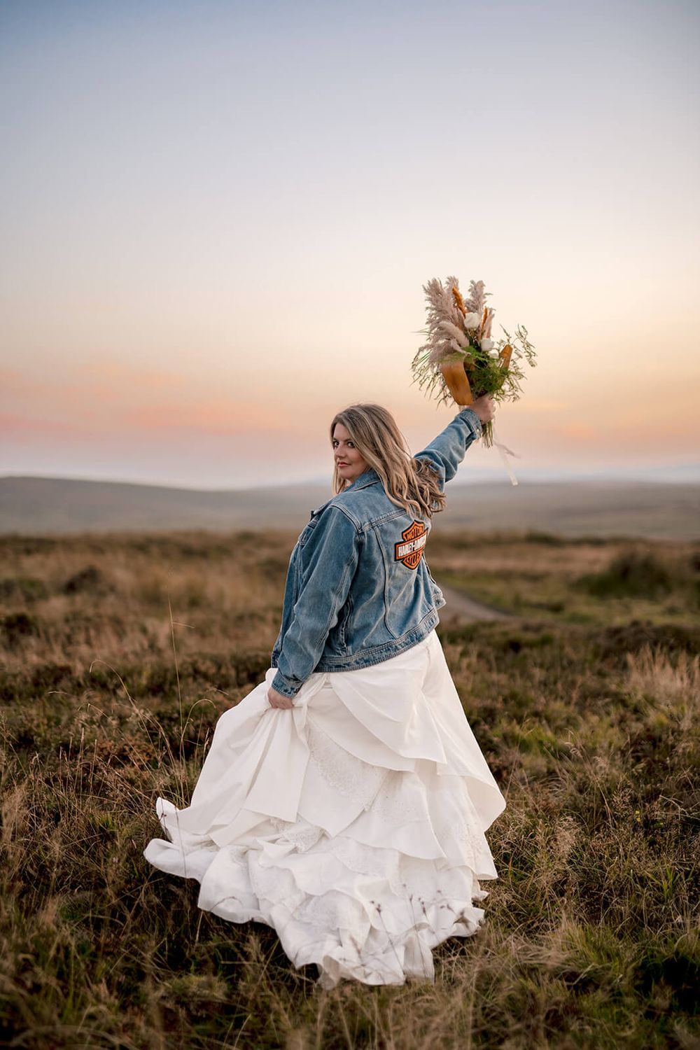 A bride in a denim jacket and flowing white dress raises her bouquet triumphantly at sunset in the mountains of a Leitrim.