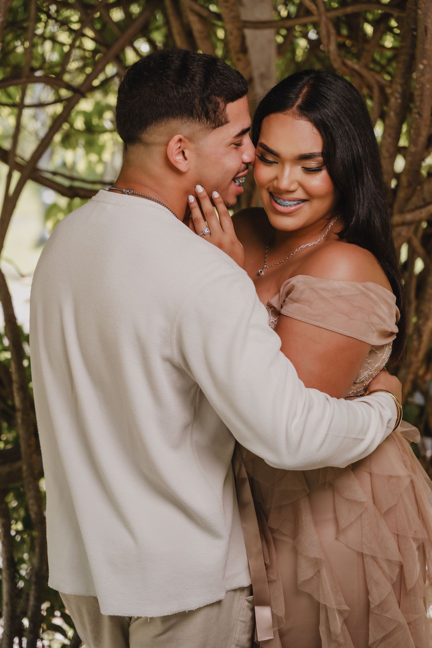 A romantic couple embraces outdoors in neutral-toned clothing, sharing an intimate moment among tree branches.