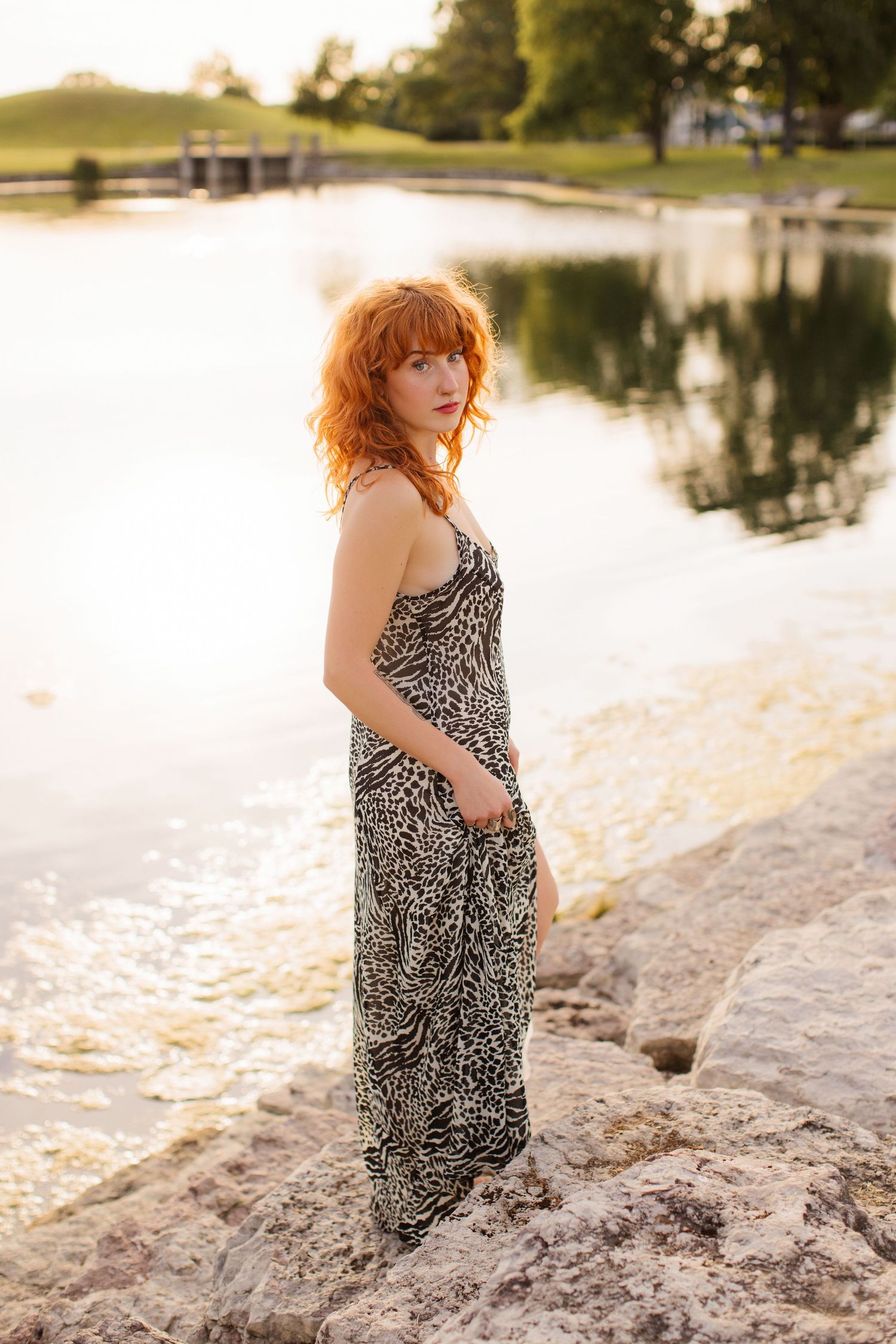 A person in a patterned dress stands on rocks beside a tranquil lake at sunset.