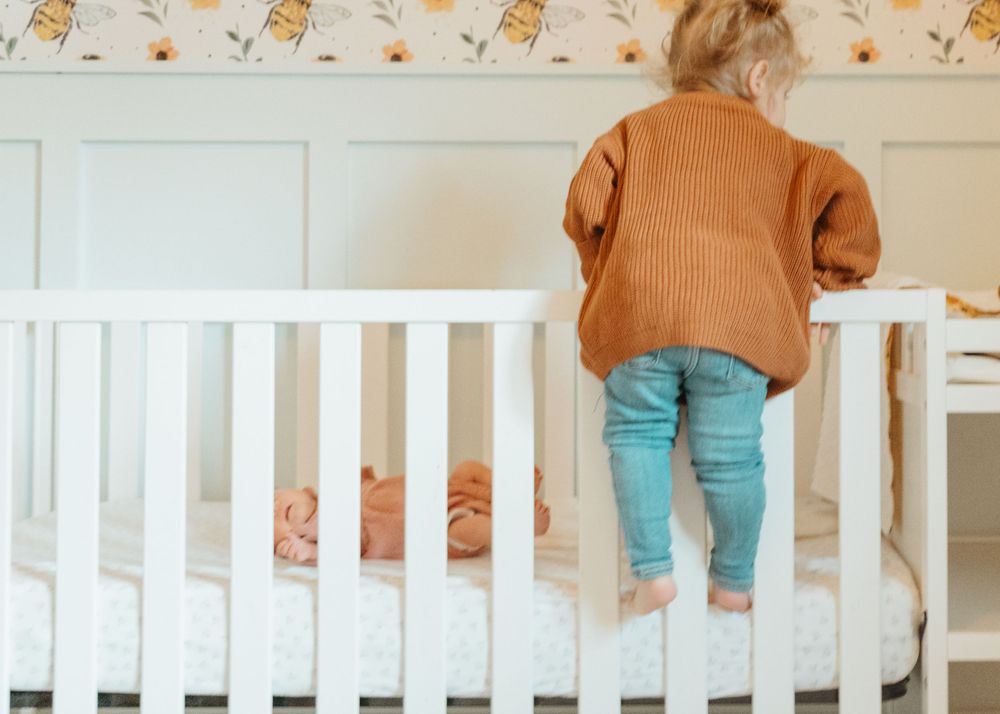Child in brown sweater standing at white crib with patterned wallpaper above.