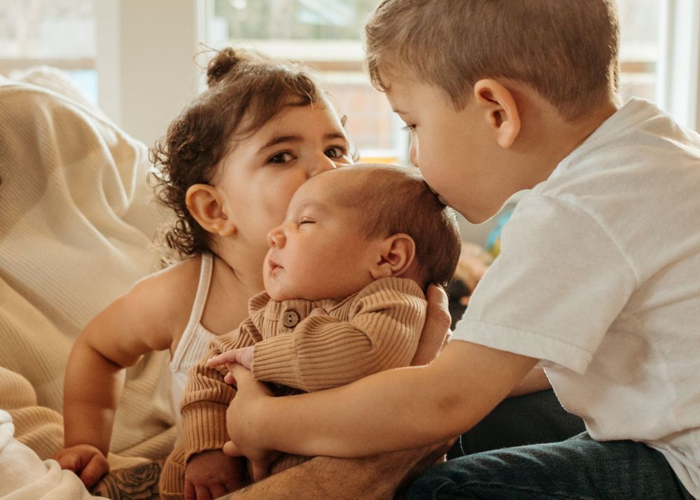 Young siblings sharing a tender moment with their baby sibling wearing a brown sweater on a light-colored couch.