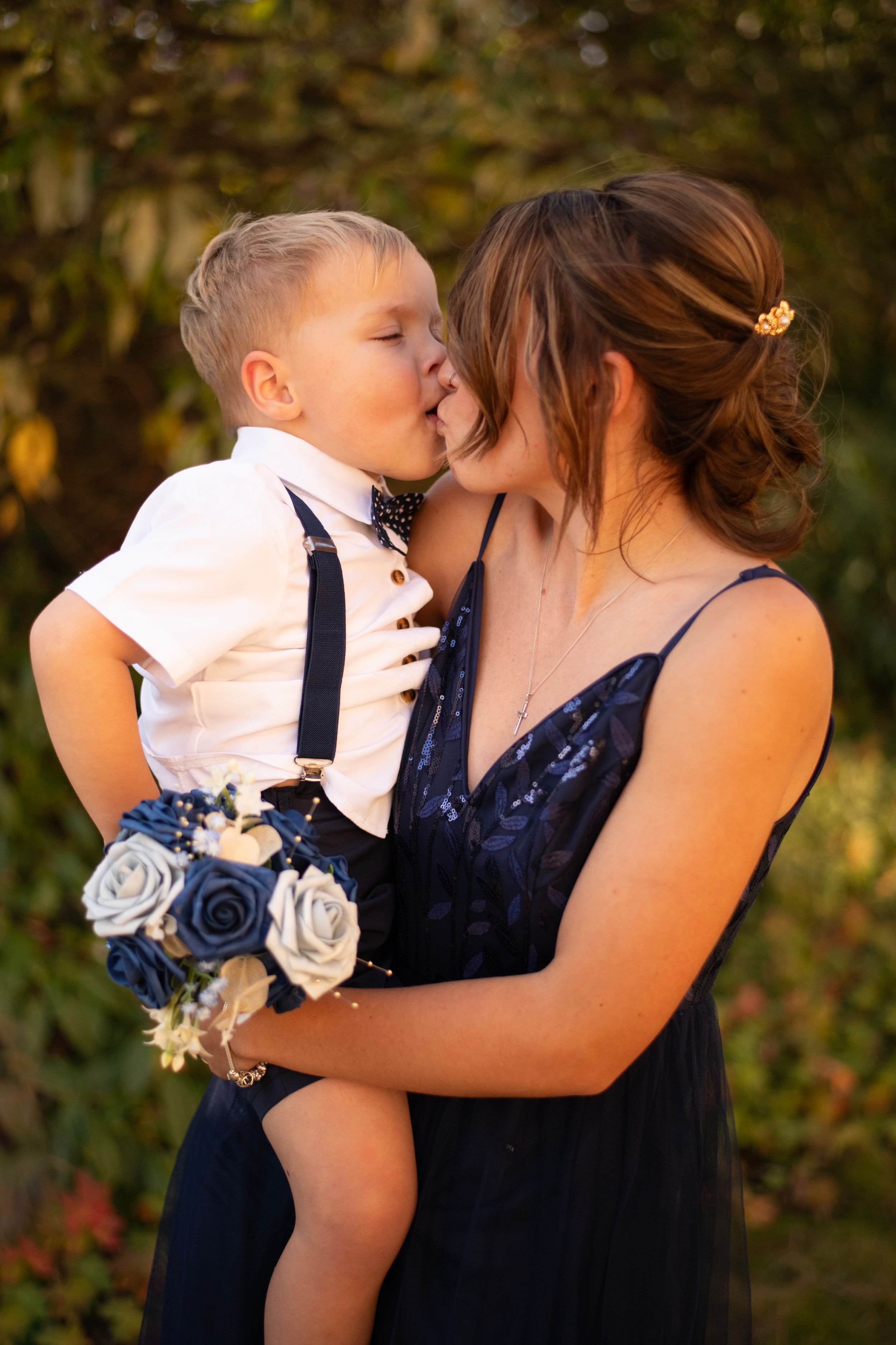 A tender moment between a young child in suspenders and a woman in a navy dress holding blue roses.
