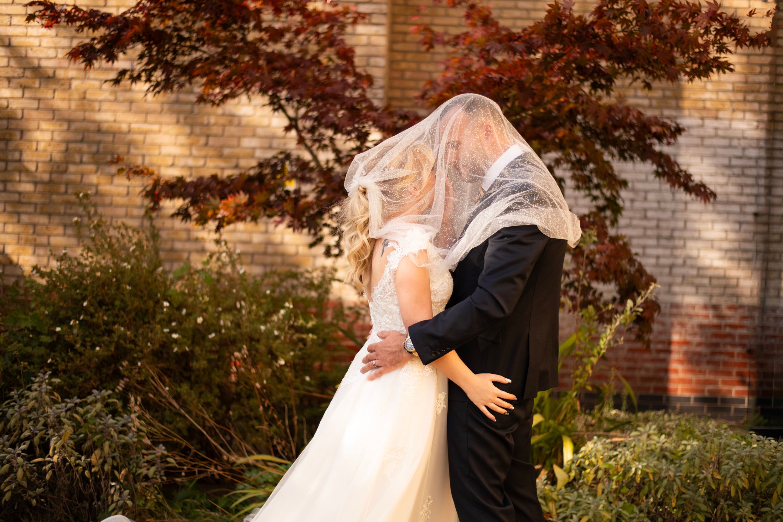 A romantic wedding photo showing a couple embracing under a veil in front of a brick building with autumn foliage.
