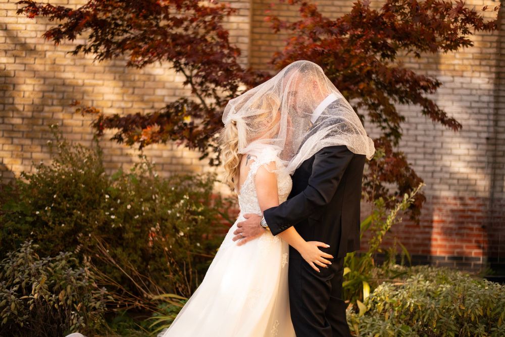 A romantic wedding photo showing a couple embracing under a veil in front of a brick building with autumn foliage.