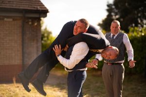 Playful wedding party members lift and dip someone in formal attire outdoors at sunset.