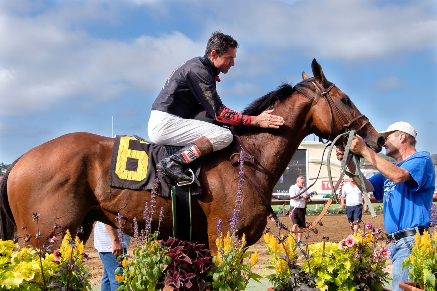 A jockey sits atop a horse while someone adjusts the saddle.