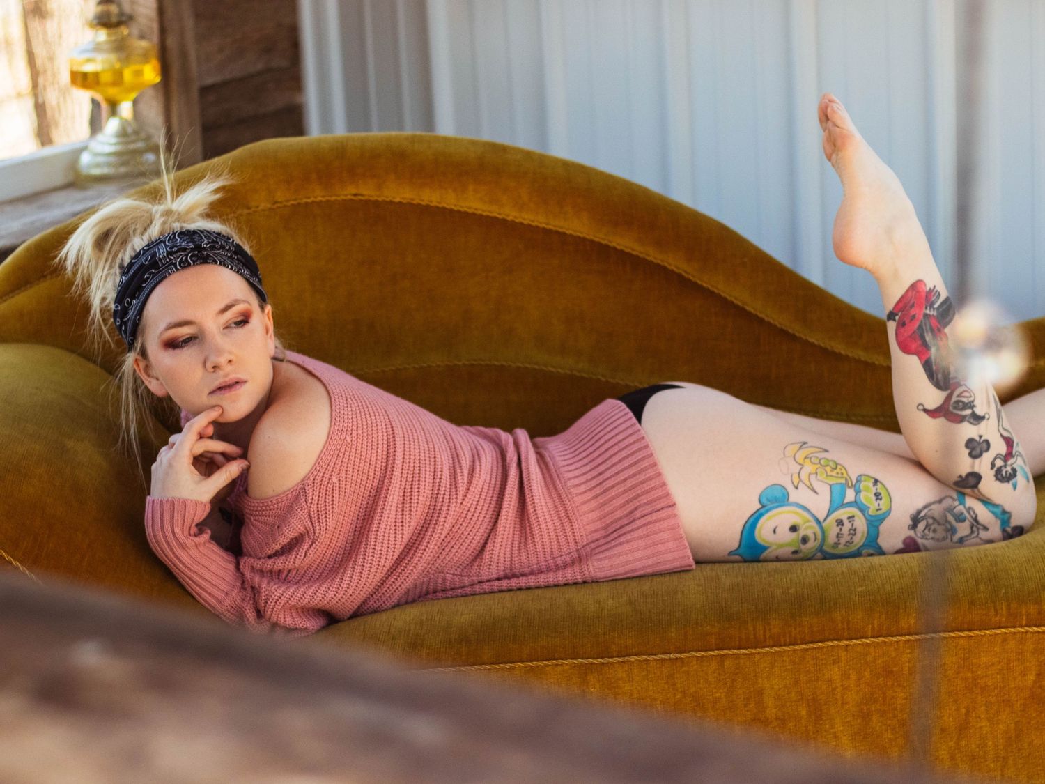 Lounging portrait on brown velvet couch showing decorative leg tattoos and pink sweater.