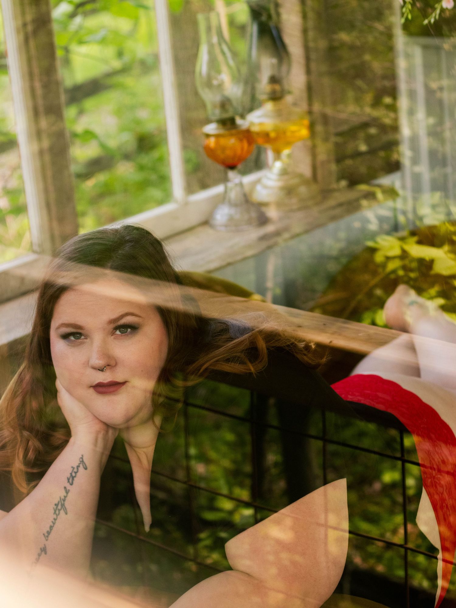 Dreamy boudoir portrait through glass with wine glasses and greenery in background.