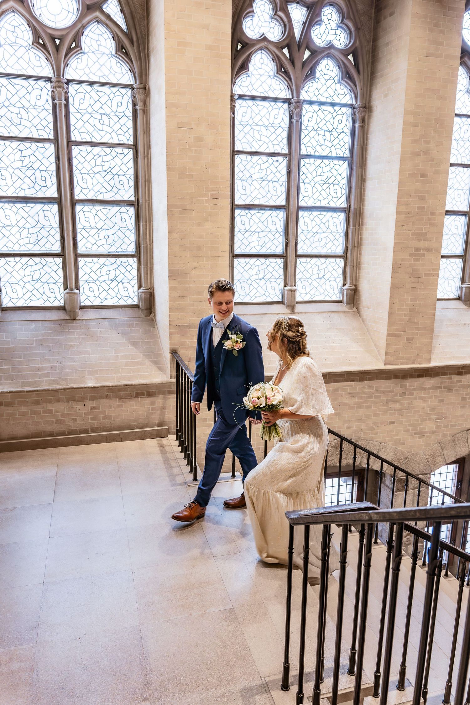 A couple walks up a grand staircase with Gothic style windows in a historic building.