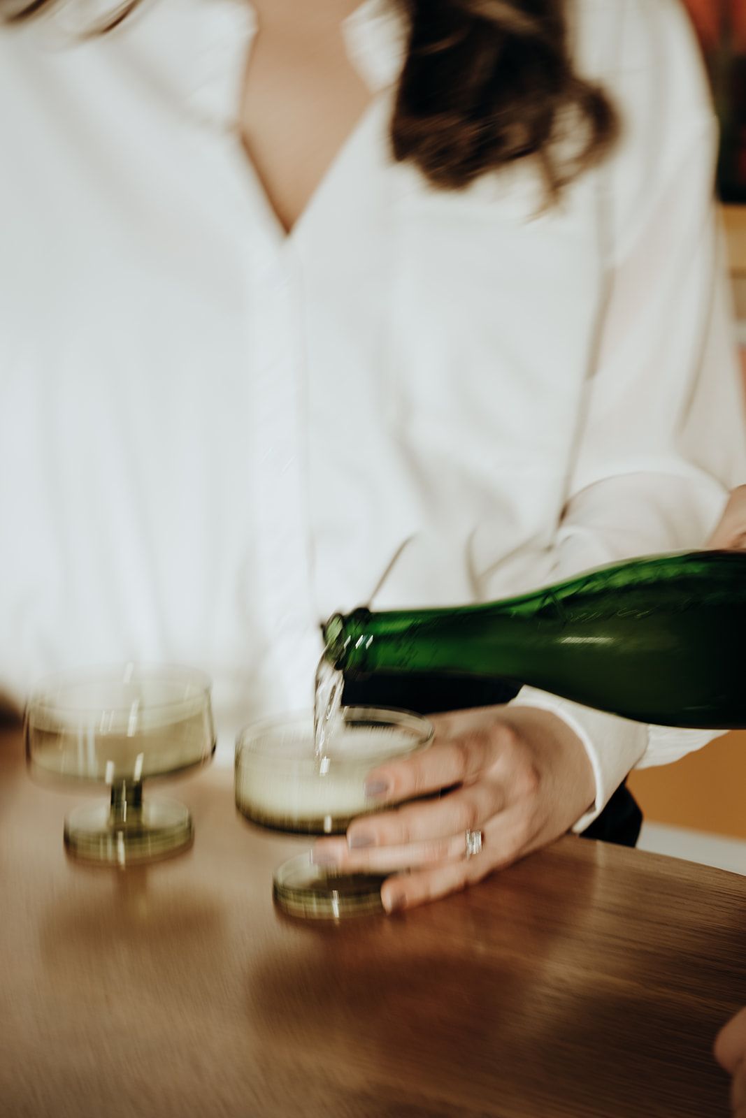Bride in white shirt pours champagne from green bottle into glasses on wooden table.