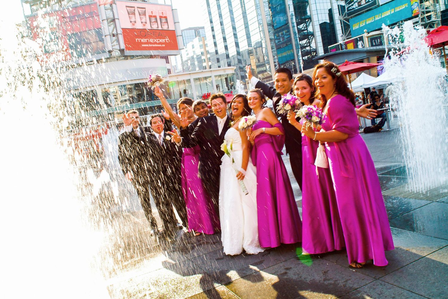 A wedding party in purple dresses poses by a fountain in an urban setting.
